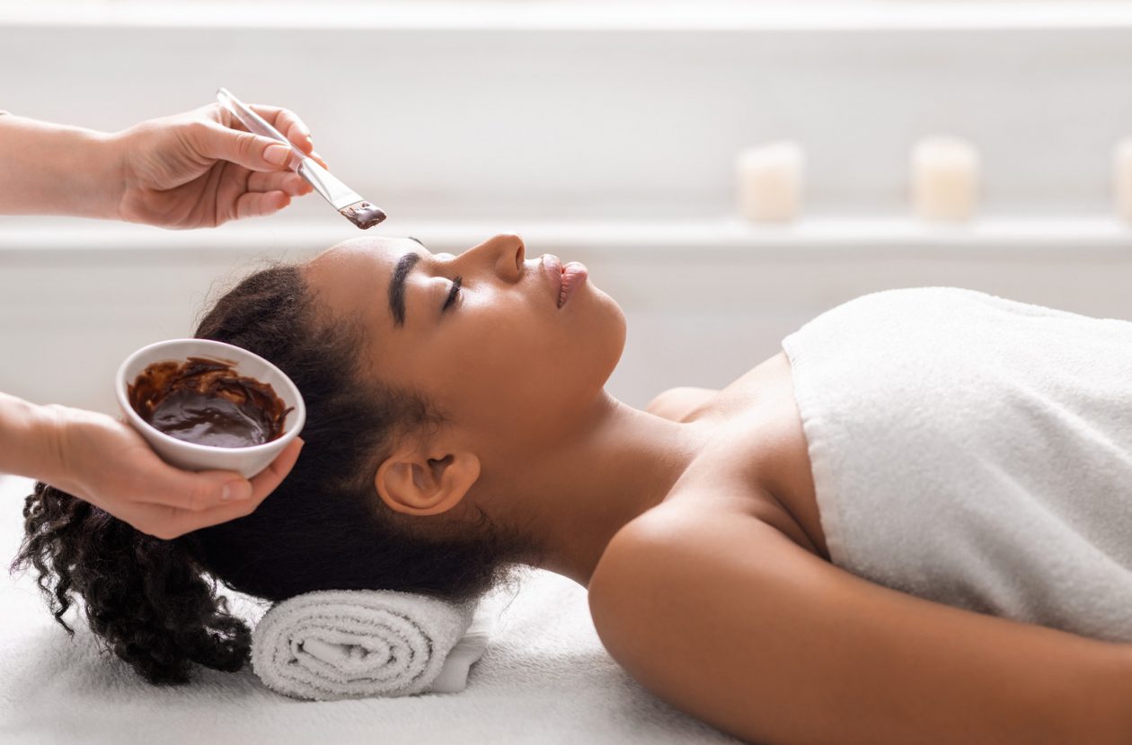 Facial treatment concept. Side view of african woman getting facial mask at spa